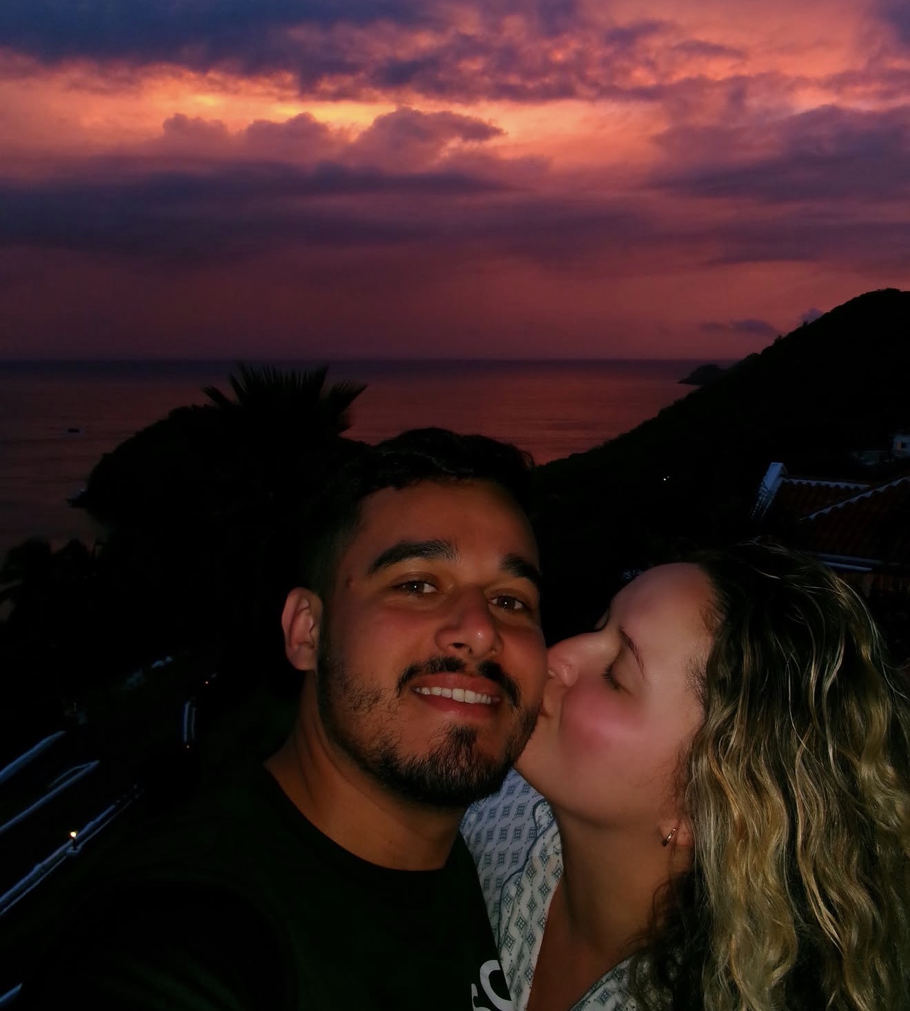 Layza kissing Matheus's cheek under a violet sunset over the sea